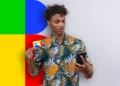 Jovem surpreso segurando cartão Bolsa Família e celular, em frente a fundo com cores primárias, ilustrando proibição no benefício