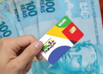 Mão segurando cartão do Programa Bolsa Família com cédulas de 100 reais ao fundo