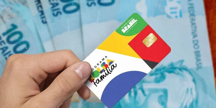 Mão segurando cartão do Programa Bolsa Família com cédulas de 100 reais ao fundo