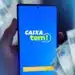 Pessoa segurando smartphone com app Caixa Tem aberto e notas de dinheiro brasileiro em destaque