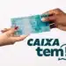 Mãos segurando uma nota de 100 reais com logo do app Caixa Tem ao fundo