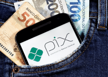 Celular com o logo do PIX ao lado de notas de real no bolso de uma calça jeans.