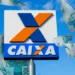 Logo da Caixa Econômica Federal com notas de 100 reais flutuando ao redor, sob um céu azul, representando liberação de fundos.