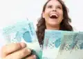 Mulher jovem e sorridente, expressando alegria, segurando várias notas de R$ 100 reais, representando o prêmio em dinheiro.