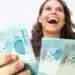 Mulher jovem e sorridente, expressando alegria, segurando várias notas de R$ 100 reais, representando o prêmio em dinheiro.