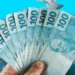 Mão segurando várias notas de 100 reais, com mais dinheiro flutuando ao fundo, simbolizando prêmios e ganhos financeiros.
