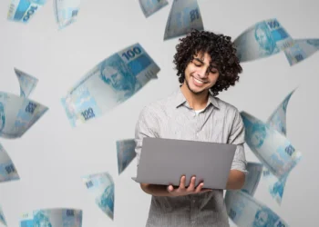 Homem jovem e sorridente com notebook, com notas de R$ 100 voando ao redor, ilustrando o sucesso ao resgatar o dinheiro esquecido em bancos pelo Sistema de Valores a Receber (SVR).