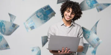 Homem jovem e sorridente com notebook, com notas de R$ 100 voando ao redor, ilustrando o sucesso ao resgatar o dinheiro esquecido em bancos pelo Sistema de Valores a Receber (SVR).