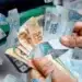 Mão segurando um leque de notas de dinheiro brasileiras (notas de 100 e 50 reais), com mais notas de 100 reais flutuando ao redor, como se estivessem caindo ou sendo distribuídas.