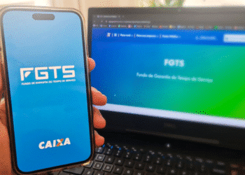 Imagem de uma mão segurando um smartphone com o aplicativo FGTS da Caixa aberto, ao lado de um laptop exibindo o site do FGTS em fundo gradiente azul e verde.