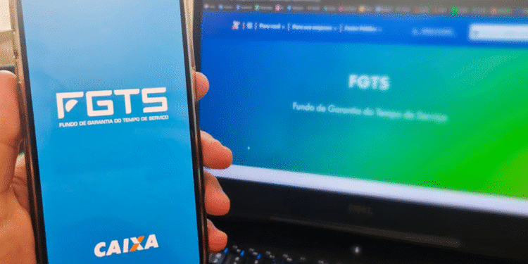 Imagem de uma mão segurando um smartphone com o aplicativo FGTS da Caixa aberto, ao lado de um laptop exibindo o site do FGTS em fundo gradiente azul e verde.