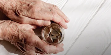 Duas mãos envelhecidas segurando um pote com moedas em uma mesa de madeira branca