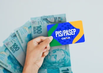 Mão segurando cartão PIS/PASEP com várias notas de 100 reais ao fundo em superfície branca