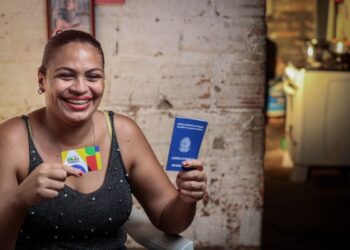 Mulher sorridente segura o cartão do Bolsa Família e sua Carteira de Trabalho, simbolizando a transição para o emprego formal com apoio do programa social.