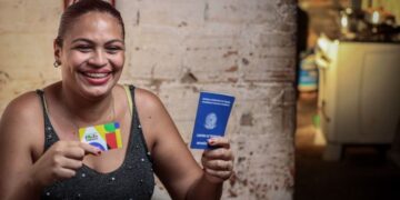Mulher sorridente segura o cartão do Bolsa Família e sua Carteira de Trabalho, simbolizando a transição para o emprego formal com apoio do programa social.