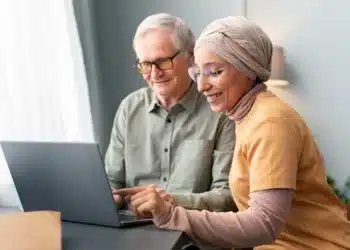 Mulher árabe ajudando homem idoso a usar laptop em ambiente doméstico, simbolizando o suporte necessário para realizar a comprovação anual de benefícios previdenciários