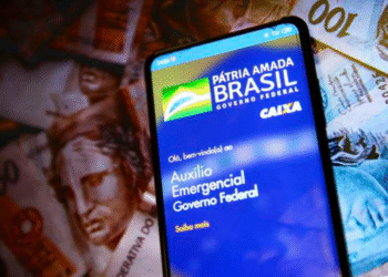 Celular exibindo a tela do aplicativo do Auxílio Emergencial do Governo Federal, com notas de real ao fundo.