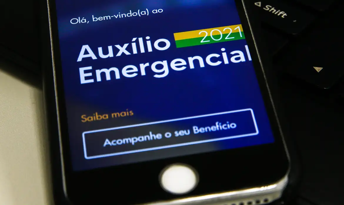 Celular com o aplicativo do Auxílio Emergencial 2021 na tela, mostrando a opção "Acompanhe o seu Benefício". Imagem simboliza a consulta para devolução dos valores recebidos indevidamente.