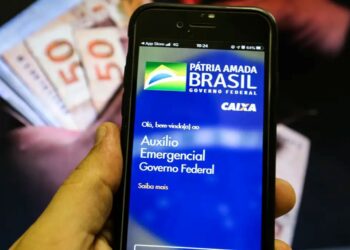 Aplicativo Auxílio Emergencial em smartphone com notas de dinheiro desfocadas ao fundo, representando a notificação para devolução.