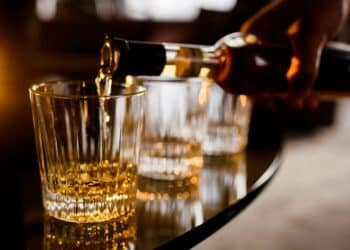 Mão de barman ou pessoa derramando uma bebida alcoólica âmbar em um copo, ilustrando o risco de intoxicação por metanol em bebidas adulteradas.