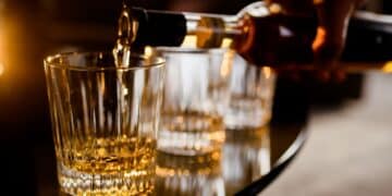 Mão de barman ou pessoa derramando uma bebida alcoólica âmbar em um copo, ilustrando o risco de intoxicação por metanol em bebidas adulteradas.