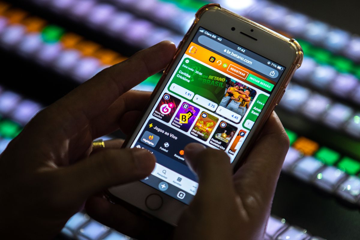 Mãos usando smartphone com aplicativo de apostas online aberto, representando a proibição para beneficiários de auxílios sociais