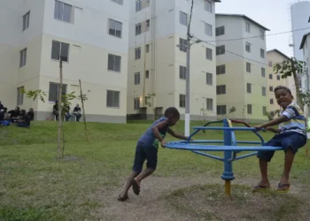 Duas crianças brincando em gira-gira em área verde de condomínio com prédios residenciais ao fundo
