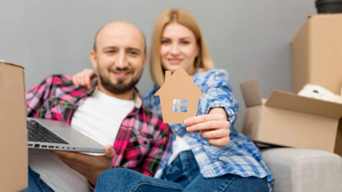 Casal sorridente sentado em um sofá com caixas de mudança, segurando um recorte de papel em formato de casa e usando um notebook para pesquisar sobre financiamento.