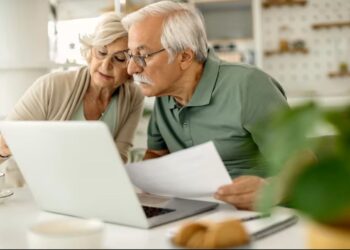 Um casal de idosos, ele com óculos e camisa polo verde, ela com óculos e blusa clara, estudando documentos e olhando para um laptop em uma mesa branca, com uma planta verde em primeiro plano.