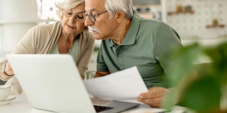 Um casal de idosos, ele com óculos e camisa polo verde, ela com óculos e blusa clara, estudando documentos e olhando para um laptop em uma mesa branca, com uma planta verde em primeiro plano.