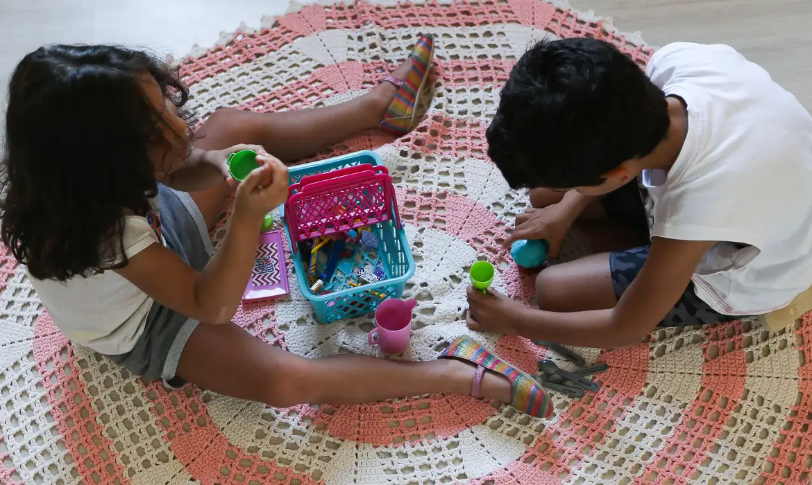 Crianças brincando com massinha em tapete de crochê Duas crianças sentadas em tapete de crochê colorido brincando com massinha e utensílios de plástico