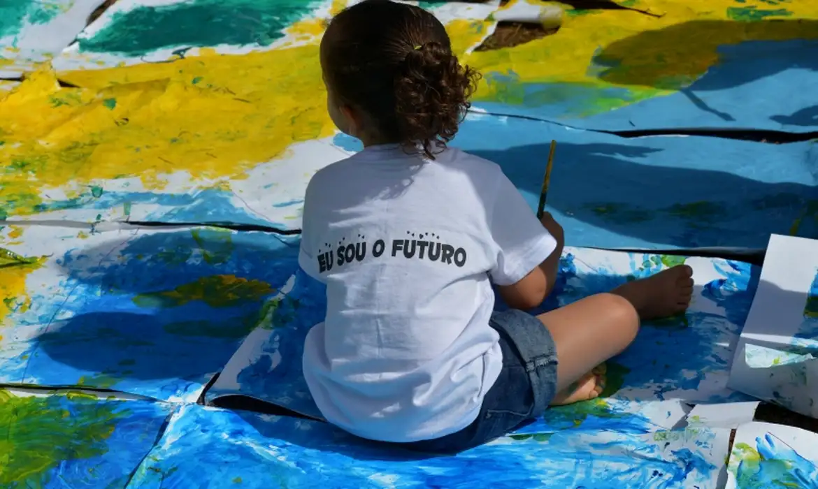 Menina sentada no chão pintando com camiseta branca escrita "Eu sou o futuro" e fundo colorido azul e amarelo
