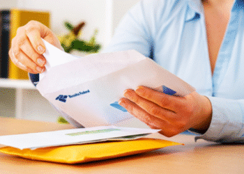 Uma pessoa com camisa azul abrindo cuidadosamente um envelope branco com o logotipo da Receita Federal em uma mesa de escritório. A imagem ilustra o momento de recebimento de uma comunicação oficial.