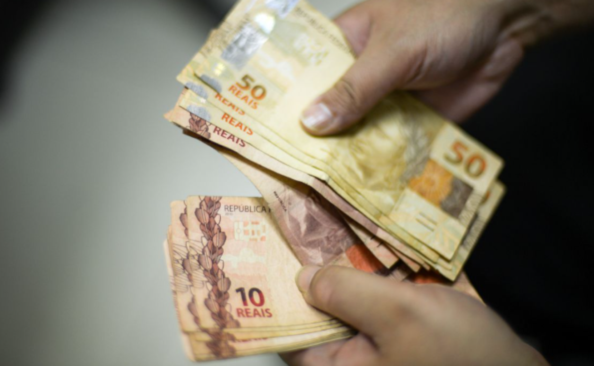 Mãos segurando notas de reais brasileiras, incluindo cédulas de 50 e 10 reais, representando pagamento ou saque de dinheiro.