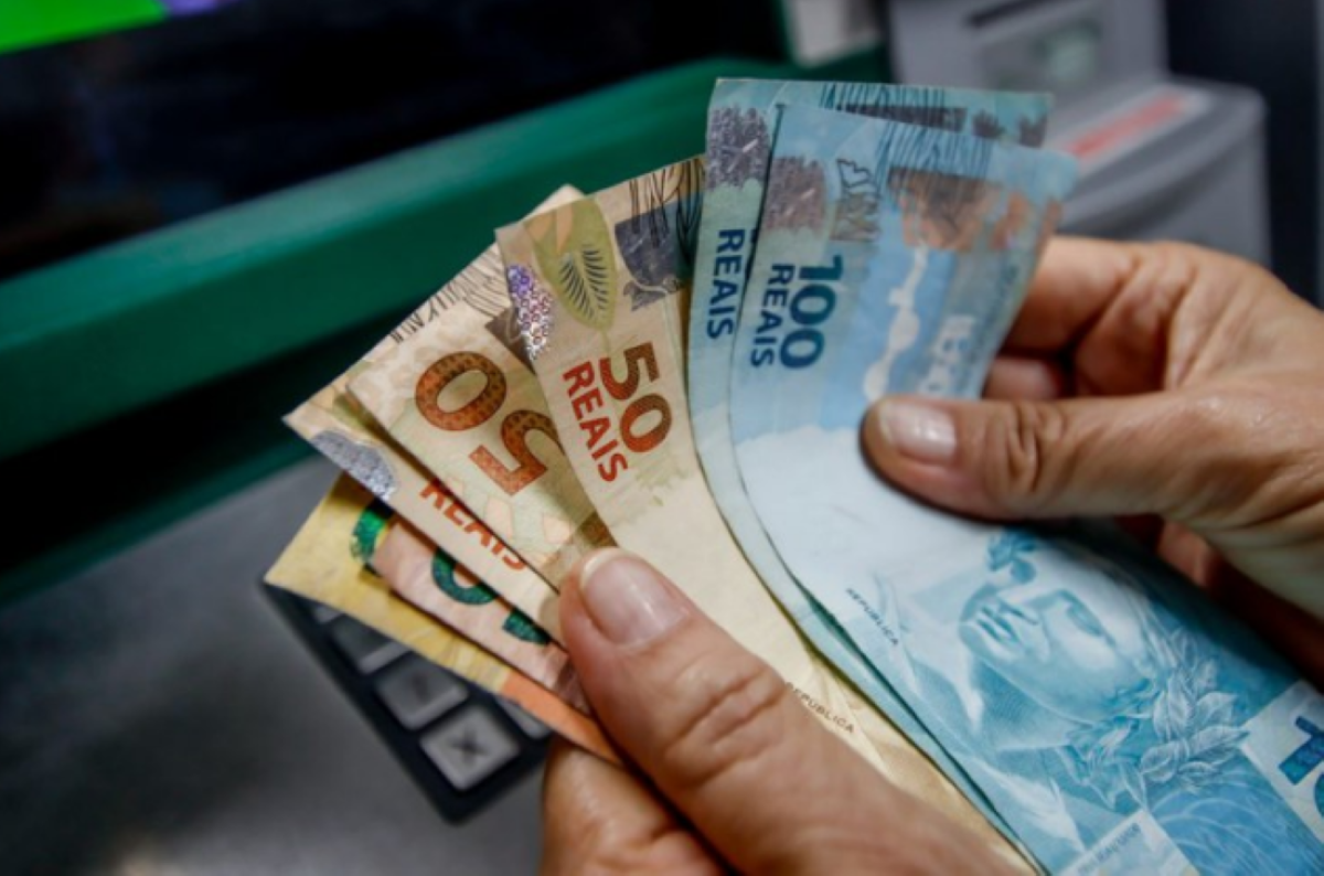 Mãos segurando cédulas de dinheiro em reais próximo a um caixa eletrônico.