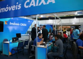Estande de atendimento da Caixa Econômica Federal em evento, com a placa "Imóveis CAIXA" e funcionários conversando com clientes.