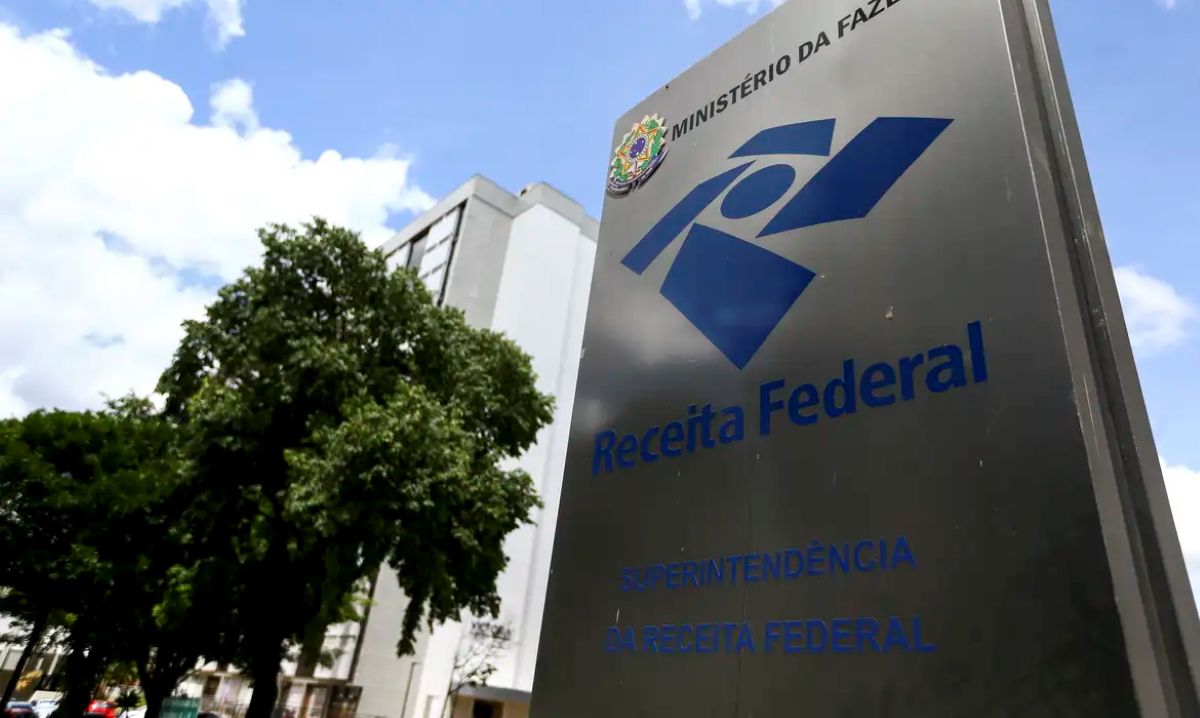 Imagem da placa cinza com o logotipo e o nome "Receita Federal", sinalizando a Superintendência do órgão, com o fundo de uma construção e árvores.