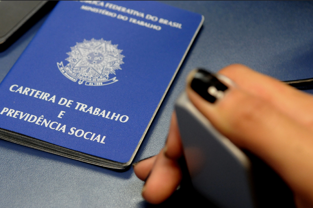 Carteira de Trabalho e Previdência Social ao lado de mão segurando celular.