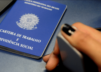 Carteira de Trabalho e Previdência Social sobre a mesa ao lado de uma mão segurando um objeto.