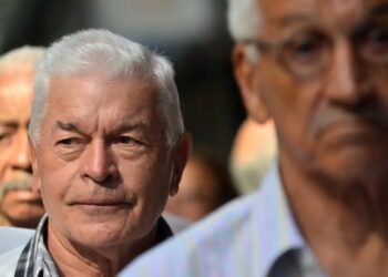 Imagem de idosos participando de uma prova de vida, com foco em um homem de cabelo branco e expressão séria, enquanto outros se encontram ao fundo.