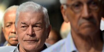 Imagem de idosos participando de uma prova de vida, com foco em um homem de cabelo branco e expressão séria, enquanto outros se encontram ao fundo.