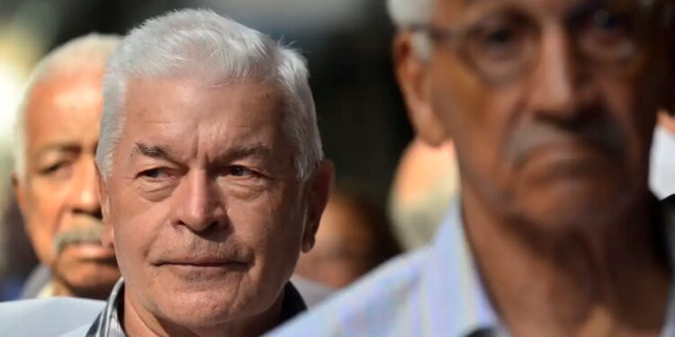 Imagem de idosos participando de uma prova de vida, com foco em um homem de cabelo branco e expressão séria, enquanto outros se encontram ao fundo.