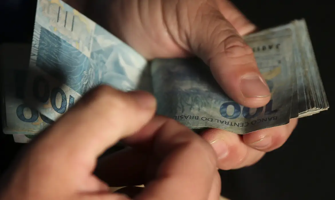 Close-up de mãos manuseando um maço de notas de R$ 100, simbolizando o dinheiro esquecido que pode ser resgatado através da consulta ao Sistema de Valores a Receber (SVR) do Banco Central.