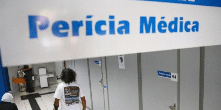 Pessoa com muleta caminhando em corredor de agência da Previdência Social em direção a salas de perícia médica, com placa indicativa no teto.