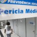 Pessoa com muleta caminhando em corredor de agência da Previdência Social em direção a salas de perícia médica, com placa indicativa no teto.