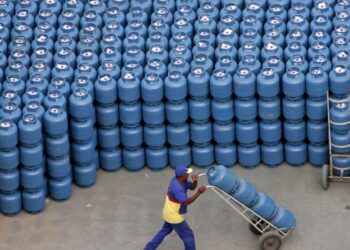 Imagem de botijões de gás empilhados, com um trabalhador transportando um deles, representando a distribuição de botijões gratuitos para 15 milhões de famílias.
