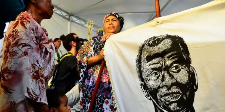 Mulheres e crianças em um evento, segurando uma bandeira com a imagem de Zumbi dos Palmares, representando a celebração e as questões do Dia da Consciência Negra.