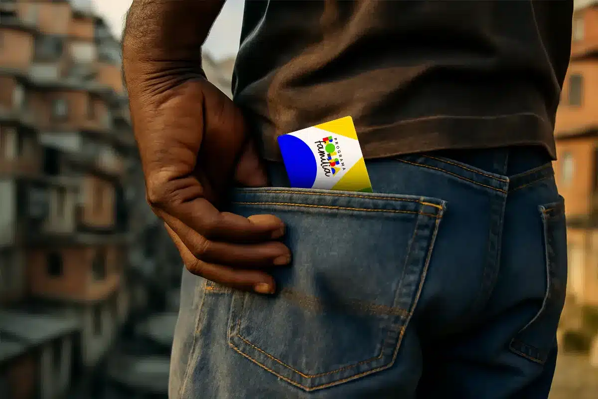 Elegibilidade Bolsa Família: Critérios para Receber Apenas R$ 300 e Como Isso Afeta Sua Família Pessoa colocando o cartão do Programa Bolsa Família no bolso traseiro de uma calça jeans, com uma comunidade ao fundo.