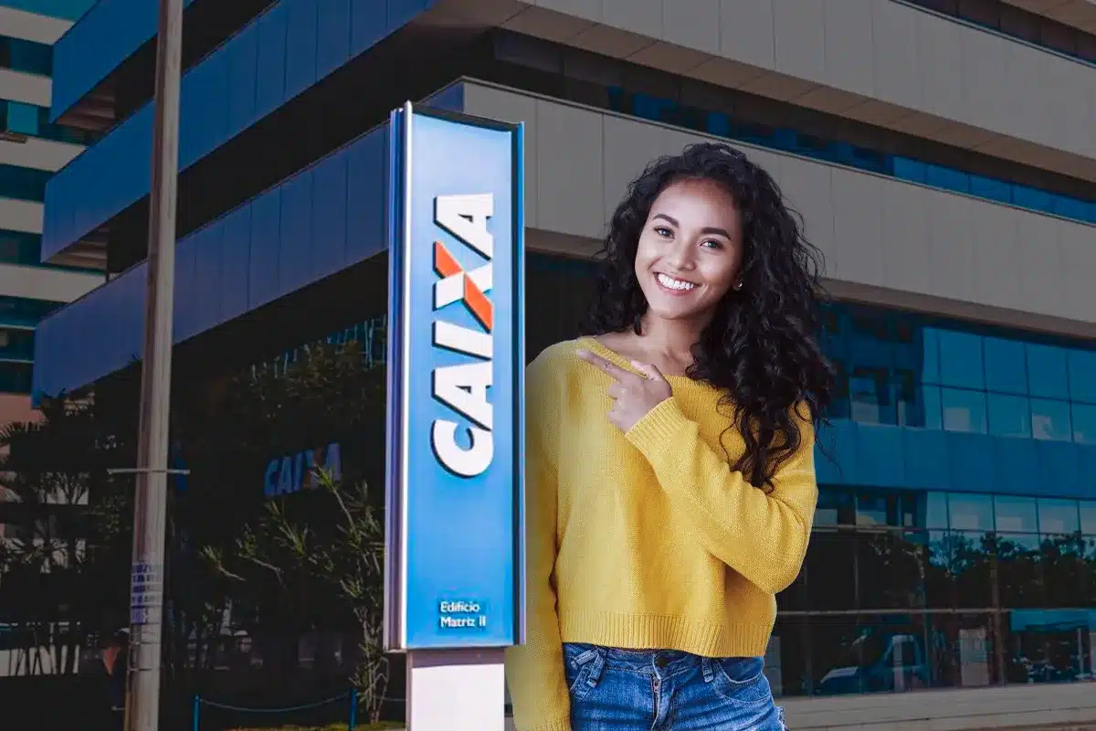 Mulher sorridente aponta para placa da Caixa Econômica Federal Mulher com suéter amarelo sorrindo e apontando para placa azul da Caixa Econômica Federal em frente a prédio moderno