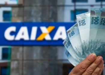 Mão segurando várias cédulas de 100 reais com logo da Caixa ao fundo desfocado